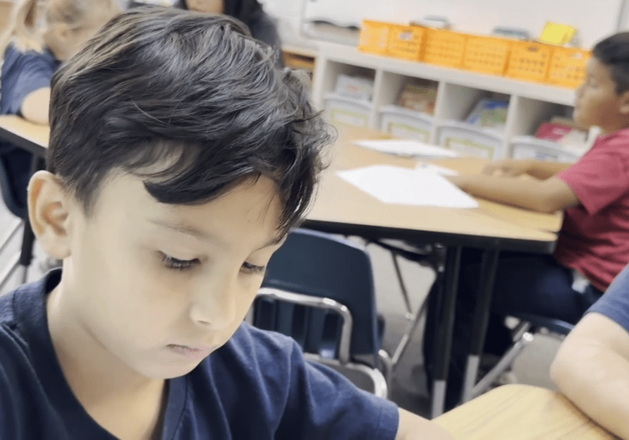 A young boy with dark hair is focused on writing or drawing at a table in a classroom setting.